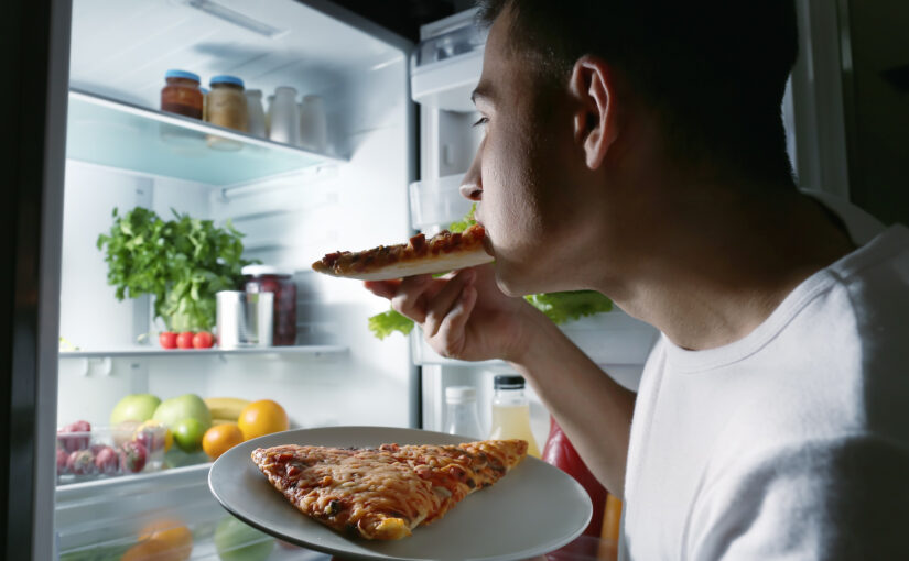 Man eating pizza in front of the fridge at night