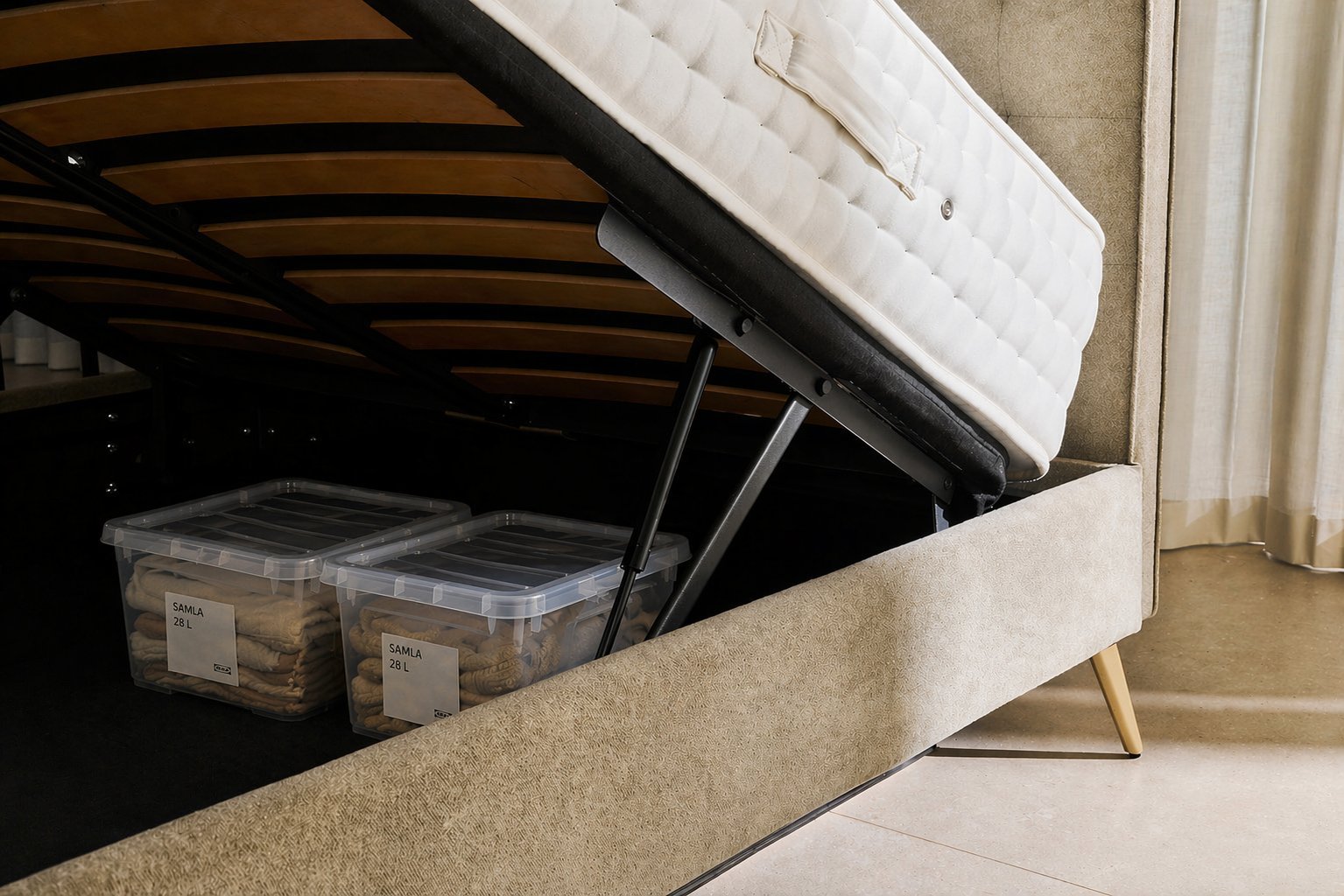 Close-up view of two labelled SAMLA 28L storage boxes standing upright under a lifted Furl ottoman bed, showing folded neutral-tone textiles inside